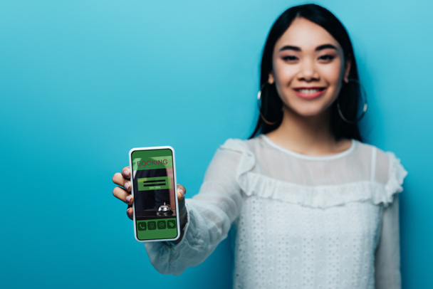 selective focus of smiling asian woman in white blouse holding smartphone with booking app on blue background - Photo, Image