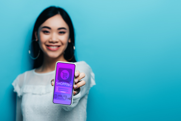 Selektiver Fokus einer lächelnden asiatischen Frau in weißer Bluse mit Smartphone mit Online-Shopping-App auf blauem Hintergrund - Foto, Bild