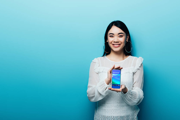 Beyaz bluzlu gülümseyen Asyalı kadın elinde telefon ile mavi arka planda yer ayırtmış. - Fotoğraf, Görsel