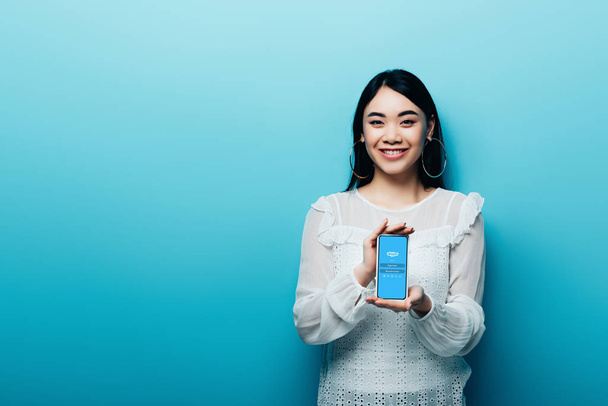 KYIV, UKRAINE - JULY 15, 2019: smiling asian woman in white blouse holding smartphone with skype app on blue background - Photo, Image