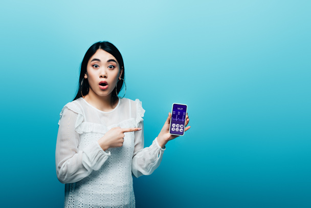 geschokt aziatische vrouw wijzen met de vinger naar smartphone met gezondheidszorg app op blauwe achtergrond - Foto, afbeelding