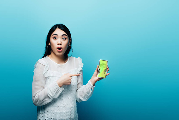 shocked asian woman pointing with finger at smartphone with fashion shopping app on blue background - Fotografie, Obrázek