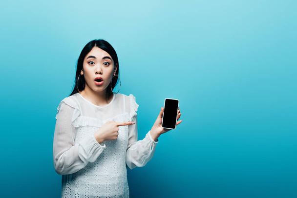 shocked asian woman pointing with finger at smartphone with blank screen on blue background - Foto, Imagen