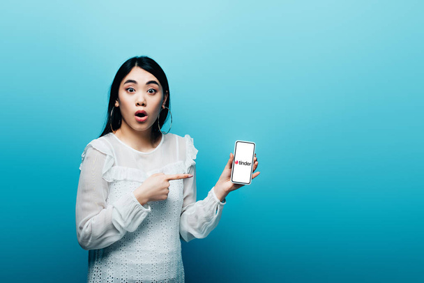 KYIV, UKRAINE - JULY 15, 2019: shocked asian woman pointing with finger at smartphone with tinder app on blue background - Фото, изображение