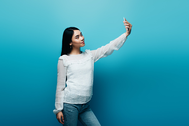 Beyaz bluzlu Asyalı kadın mavi arka planda akıllı telefondan selfie çekiyor. - Fotoğraf, Görsel