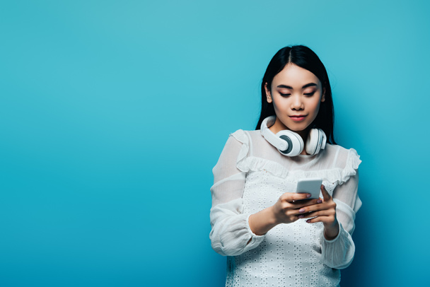 asian woman with wireless headphones in white blouse using smartphone on blue background - Fotó, kép
