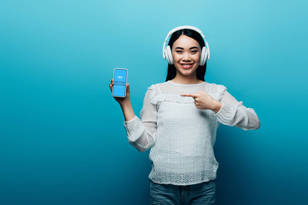 KYIV, UKRAINE - JULY 15, 2019: smiling asian woman with headphones pointing with finger at smartphone with skype icon on blue background - Fotó, kép