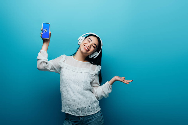 KYIV, UKRAINE - JULY 15, 2019: smiling asian woman with closed eyes in headphones holding smartphone with Shazam app and dancing on blue background - Zdjęcie, obraz