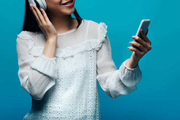 cropped view of smiling asian woman with headphones using smartphone on blue background - Zdjęcie, obraz
