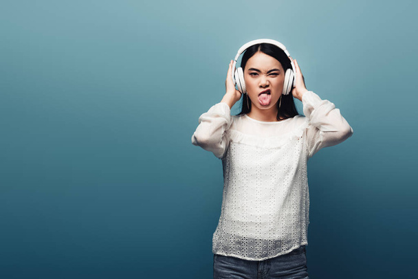 asiatische Frau mit Kopfhörer Grimassen auf blauem Hintergrund - Foto, Bild