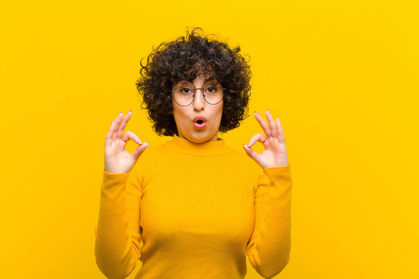 young pretty afro woman feeling shocked, amazed and surprised, showing approval making okay sign with both hands - Фото, зображення