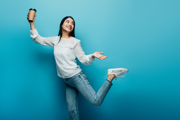 happy brunette asian woman posing with coffee to go on blue background - Foto, Imagem