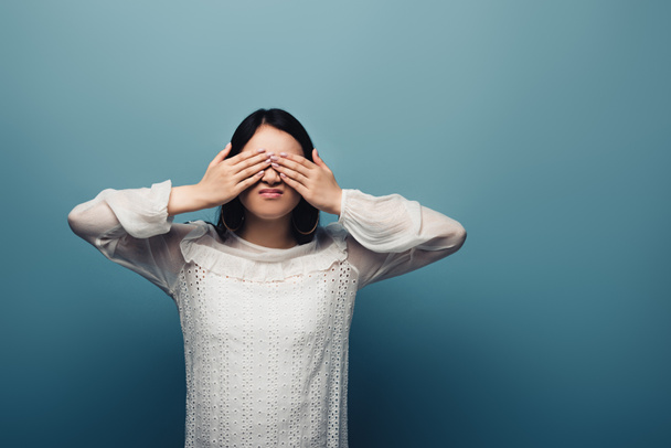 upset brunette asian woman with hands on eyes on blue background - Photo, image
