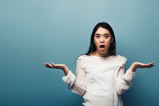 shocked brunette asian woman showing shrug gesture on blue background - Foto, immagini