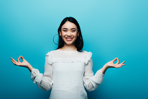 happy brunette asian woman meditating on blue background - Zdjęcie, obraz