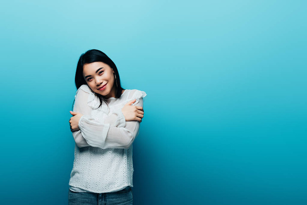 happy brunette asian woman hugging herself on blue background - Photo, Image