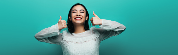 happy brunette asian woman showing thumbs up on turquoise background, panoramic shot - Photo, Image