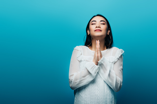 брюнетка азіатська жінка з закритими очима і молитовними руками на синьому фоні
 - Фото, зображення