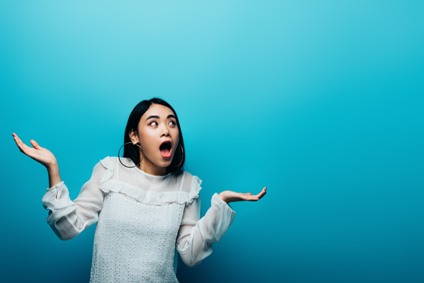 excited brunette asian woman with open mouth showing shrug gesture on blue background - Valokuva, kuva
