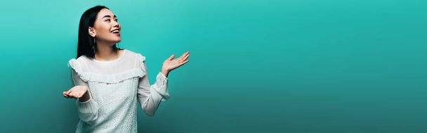 happy brunette asian woman showing shrug gesture on turquoise background, panoramic shot - Photo, Image