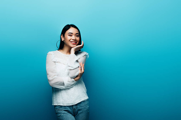 happy brunette asian woman with hand near face on blue background - Fotó, kép