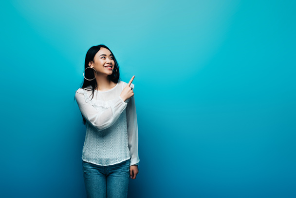 happy brunette asian woman pointing with finger on blue background - Photo, image
