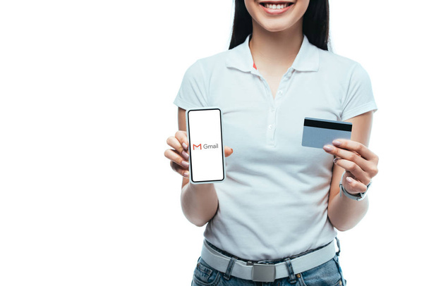 KYIV, UKRAINE - JULY 15, 2019: cropped view of smiling brunette asian girl holding credit card and smartphone with gmail app isolated on white - Fotografie, Obrázek