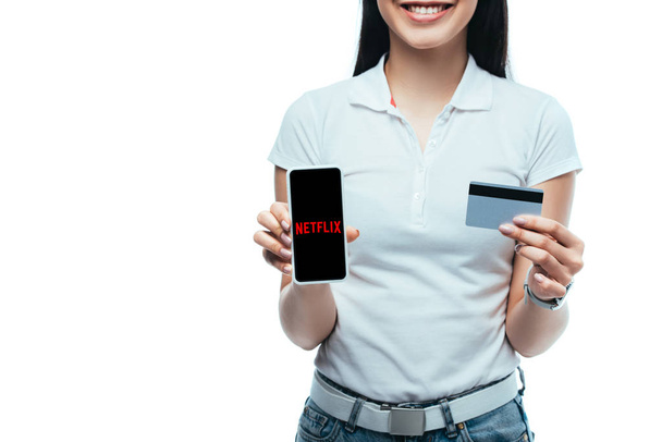 KYIV, UKRAINE - JULY 15, 2019: cropped view of smiling brunette asian girl holding credit card and smartphone with netflix app isolated on white - Фото, изображение