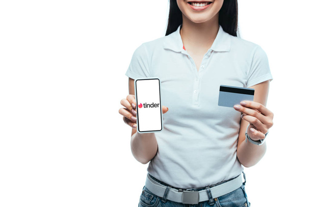 KYIV, UKRAINE - JULY 15, 2019: cropped view of smiling brunette asian girl holding credit card and smartphone with tinder app isolated on white - Foto, afbeelding