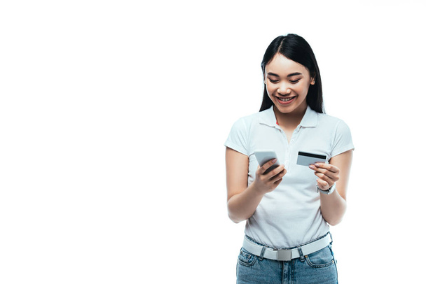 happy brunette asian girl holding credit card and smartphone isolated on white - Zdjęcie, obraz