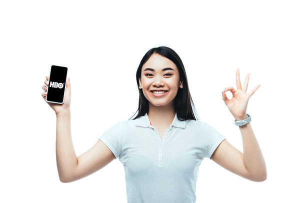 Kyiv,ウクライナ- 2019年7月15日:幸せなブルネットアジアの女性がHboアプリでスマートフォンを保持し、白い上に隔離されたokのサインを表示 - 写真・画像