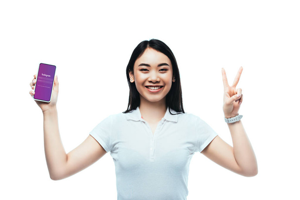 KYIV, UKRAINE HEINÄKUU 15, 2019: onnellinen brunette aasialainen nainen, jolla on älypuhelin Instagram-sovelluksella ja joka näyttää rauhan merkin eristettynä valkoisella
 - Valokuva, kuva