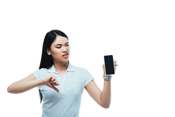 nespokojená brunetka asijské žena drží smartphone s prázdnou obrazovkou a ukazující palec dolů izolované na bílé - Fotografie, Obrázek