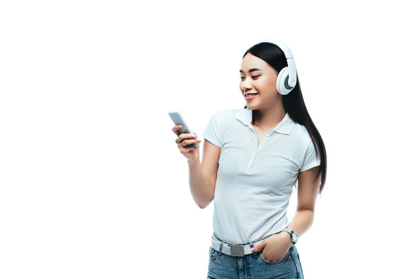 χαμογελαστό ελκυστικό κορίτσι από την Ασία με ακουστικά χρησιμοποιώντας smartphone απομονωμένο σε λευκό - Φωτογραφία, εικόνα