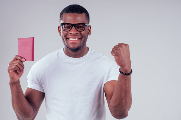 African american man student in a white T-shirt and glasses holding passport red cover - Фото, изображение