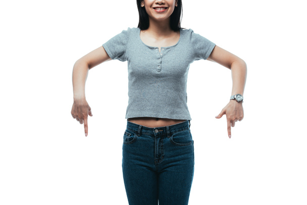abgeschnittene Ansicht eines lächelnden attraktiven asiatischen Mädchens, das mit den Fingern nach unten zeigt, isoliert auf weißem Grund - Foto, Bild