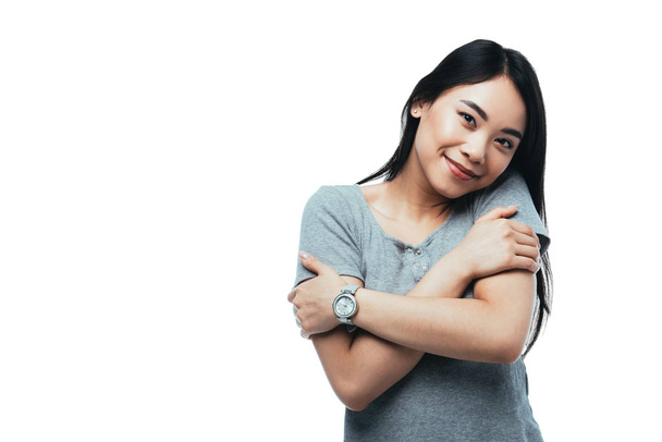 Улыбающаяся привлекательная азиатская девушка, обнимающая себя изолированной на белом
 - Фото, изображение