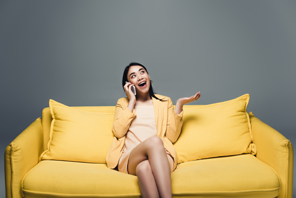 šťastná asijská podnikatelka sedí na žluté pohovce se zkříženýma nohama a mluví na smartphone na šedém pozadí - Fotografie, Obrázek