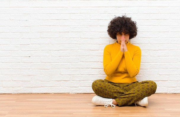 молодая красивая афро женщина чувствует себя обеспокоенной, надежной и религиозной, преданно молясь с отжатыми ладонями, умоляя о прощении сидя на полу
 - Фото, изображение
