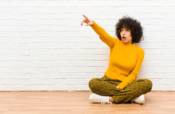 young pretty afro woman feeling shocked and surprised, pointing and looking upwards in awe with amazed, open-mouthed look sitting on the floor - 写真・画像