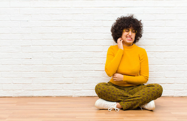 young pretty afro woman feeling stressed, frustrated and tired, rubbing painful neck, with a worried, troubled look sitting on the floor - 写真・画像