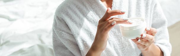 panoramic shot of woman in bathrobe holding cosmetic cream at morning  - Foto, afbeelding