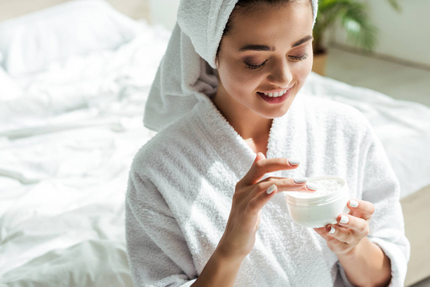 attractive woman in bathrobe and towel smiling and holding cosmetic cream at morning  - Photo, Image