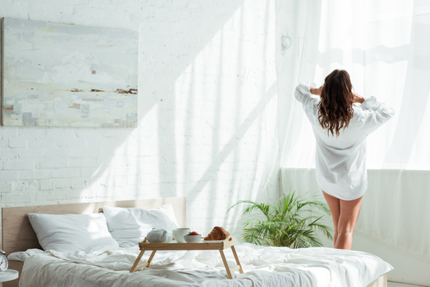 Rückansicht einer Frau im weißen Hemd, die morgens durch das Fenster schaut  - Foto, Bild