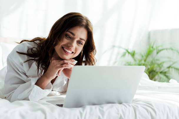 viehättävä nainen valkoisessa paidassa kannettavan tietokoneen avulla ja hymyillen aamulla
  - Valokuva, kuva