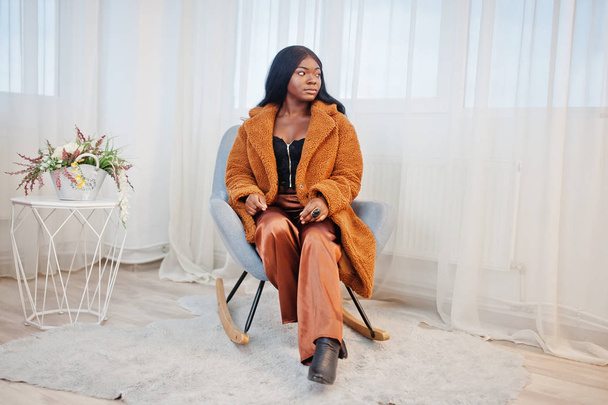 Stylish african american woman in orange coat posed at room sitt - 写真・画像