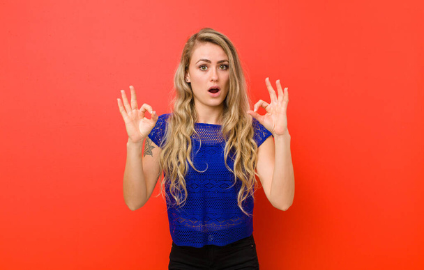 young blonde woman feeling shocked, amazed and surprised, showing approval making okay sign with both hands against red wall - Foto, immagini