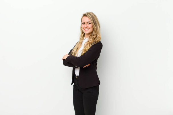 young blonde woman smiling to camera with crossed arms and a happy, confident, satisfied expression, lateral view against white wall - Фото, изображение