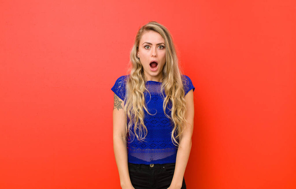 young blonde woman feeling terrified and shocked, with mouth wide open in surprise against red wall - Foto, immagini