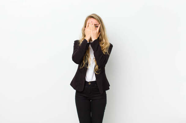 young blonde woman covering face with hands, peeking between fingers with surprised expression and looking to the side against white wall - Φωτογραφία, εικόνα